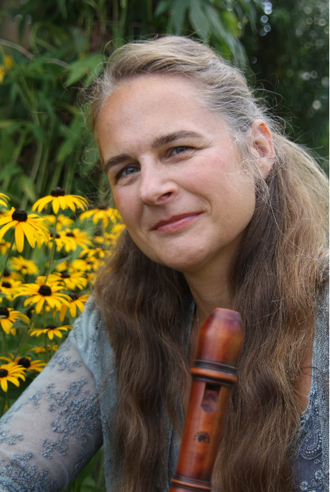 AI generated: The image shows a woman sitting in a garden with yellow flowers and holding a wooden wind instrument. She is smiling at the camera and green plants are visible in the background.