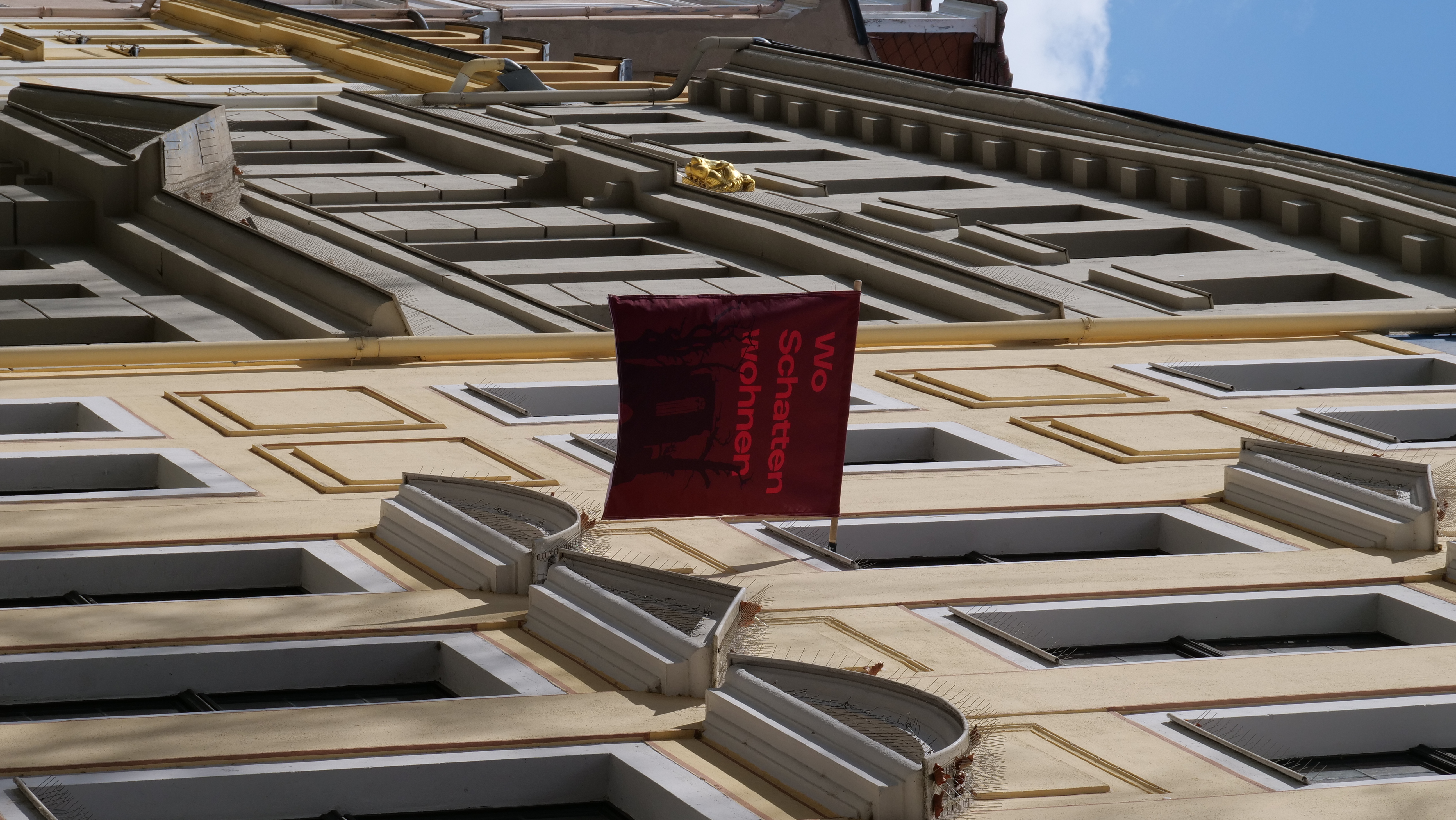 KI generiert: Das Bild zeigt die Fassade eines Gebäudes mit einer roten Fahne, auf der "Wo Schatten wohnen" steht. Die Architektur hat gelbe und beige Verzierungen und die Szene wird von Sonnenlicht beleuchtet.