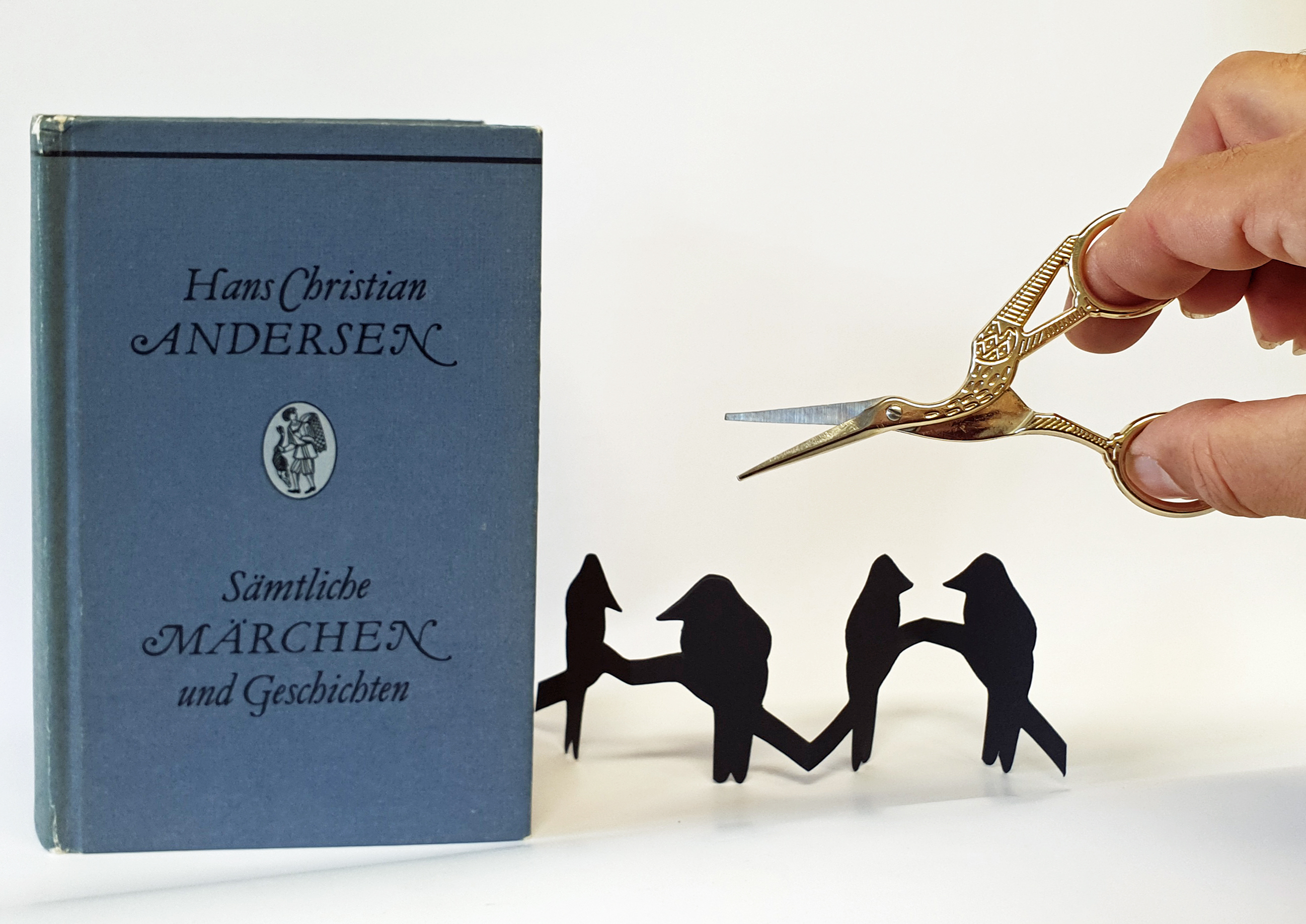 KI generiert: Ein Buch von Hans Christian Andersen steht neben einer Hand, die eine kleine Schere, geformt als Storch, hält. Dazwischen ist ein ausgeschnittenes schwarzes Papier zu sehen, das Figuren darstellt.