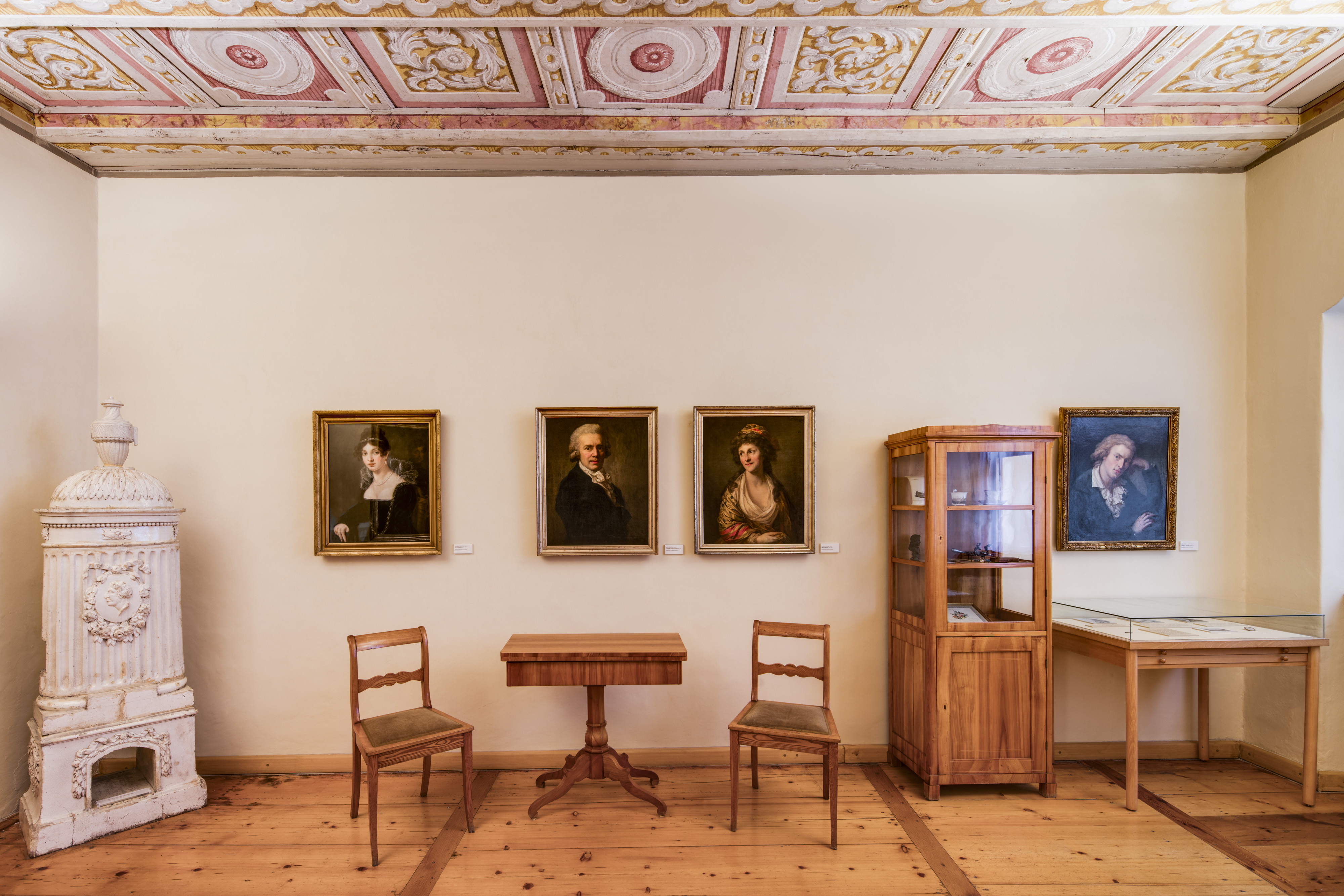 KI generiert: Das Bild zeigt einen Raum mit klassischer Einrichtung, darunter ein weißer Ofen, ein Holztisch mit zwei Stühlen, eine Vitrine und mehrere Porträtgemälde an der Wand. Die Decke ist kunstvoll mit barocken Mustern verziert.
