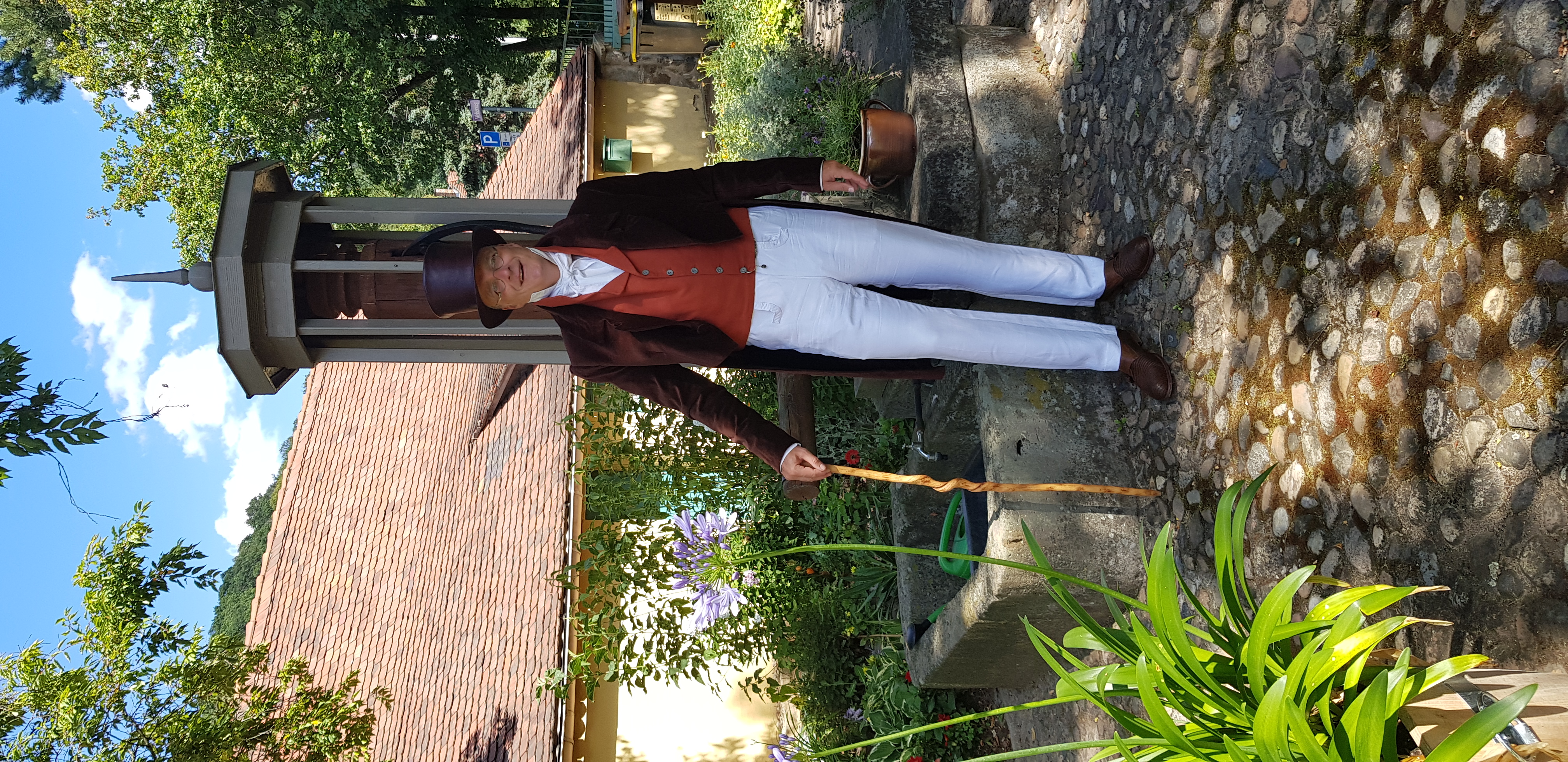 KI generiert: Ein Mann steht in historischer Kleidung mit Zylinder und Spazierstock neben einem kleinen Brunnen in einer Gartenumgebung. Im Hintergrund sind ein Gebäude mit Ziegeldach und grüne Pflanzen zu sehen.