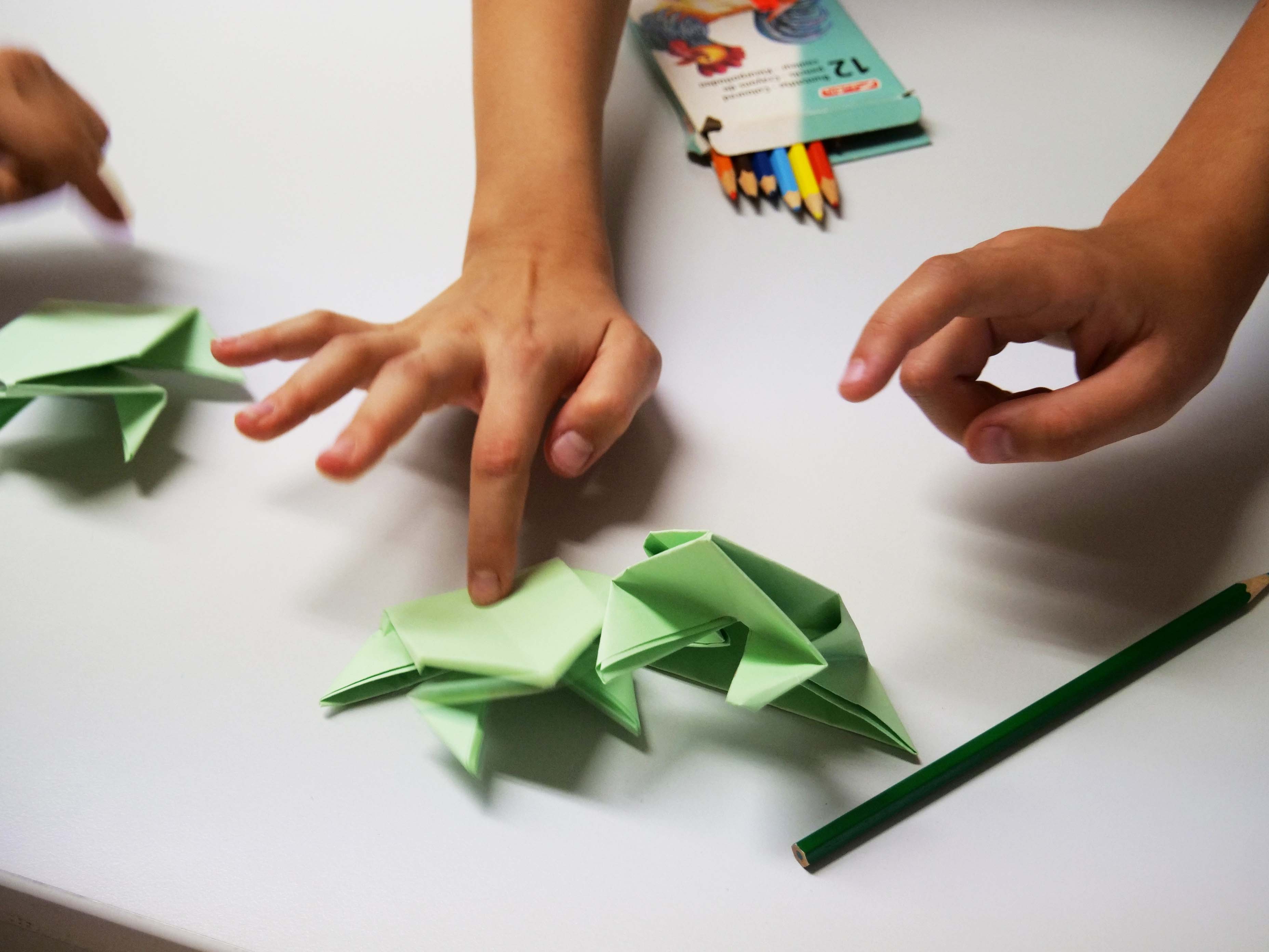 KI generiert: Das Bild zeigt Hände, die sich mit Origami beschäftigen, wobei mehrere grüne Papierfrösche zu sehen sind. Im Hintergrund sind Buntstifte und ein Buch erkennbar.