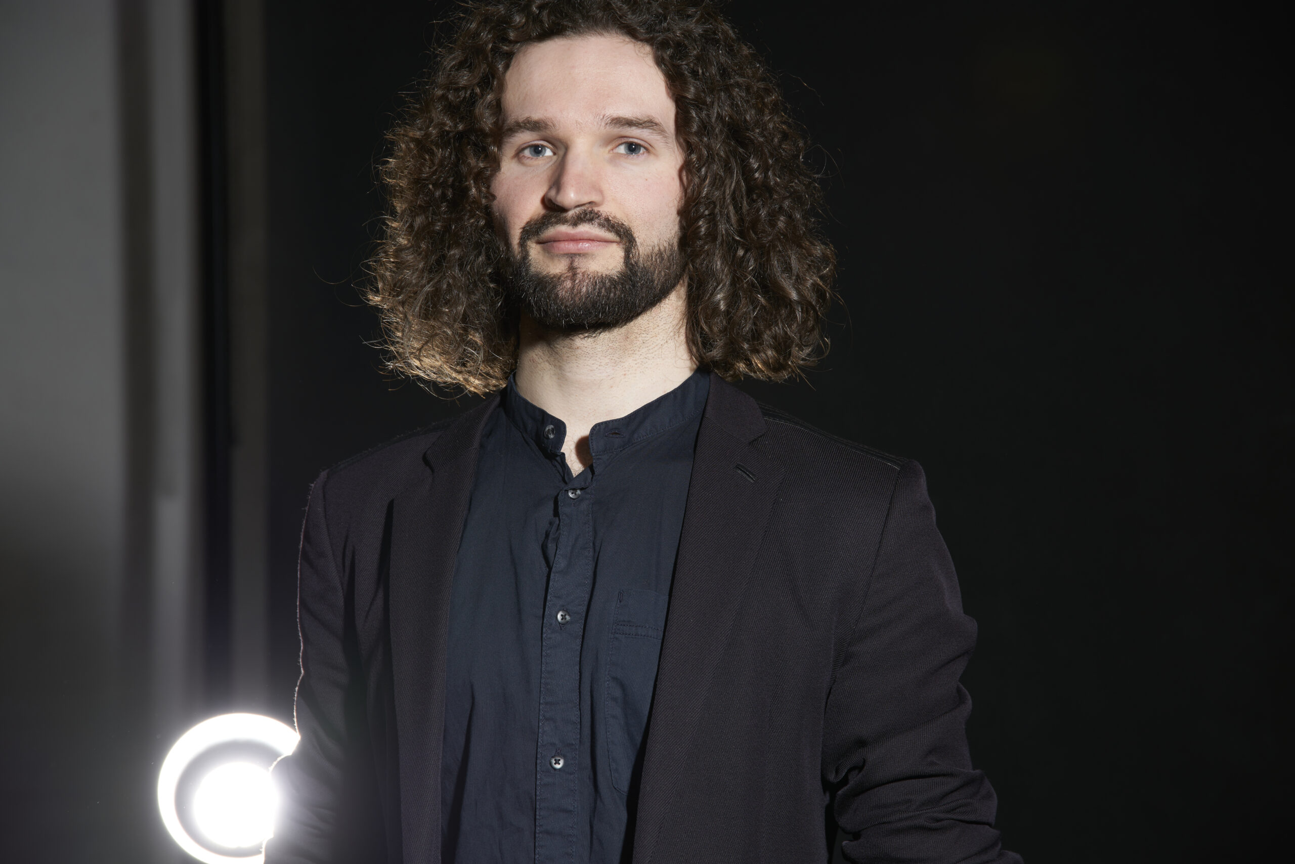 AI generated: A man with curly hair and a beard stands in front of a dark background and wears a dark suit. A round light shines at the bottom left.