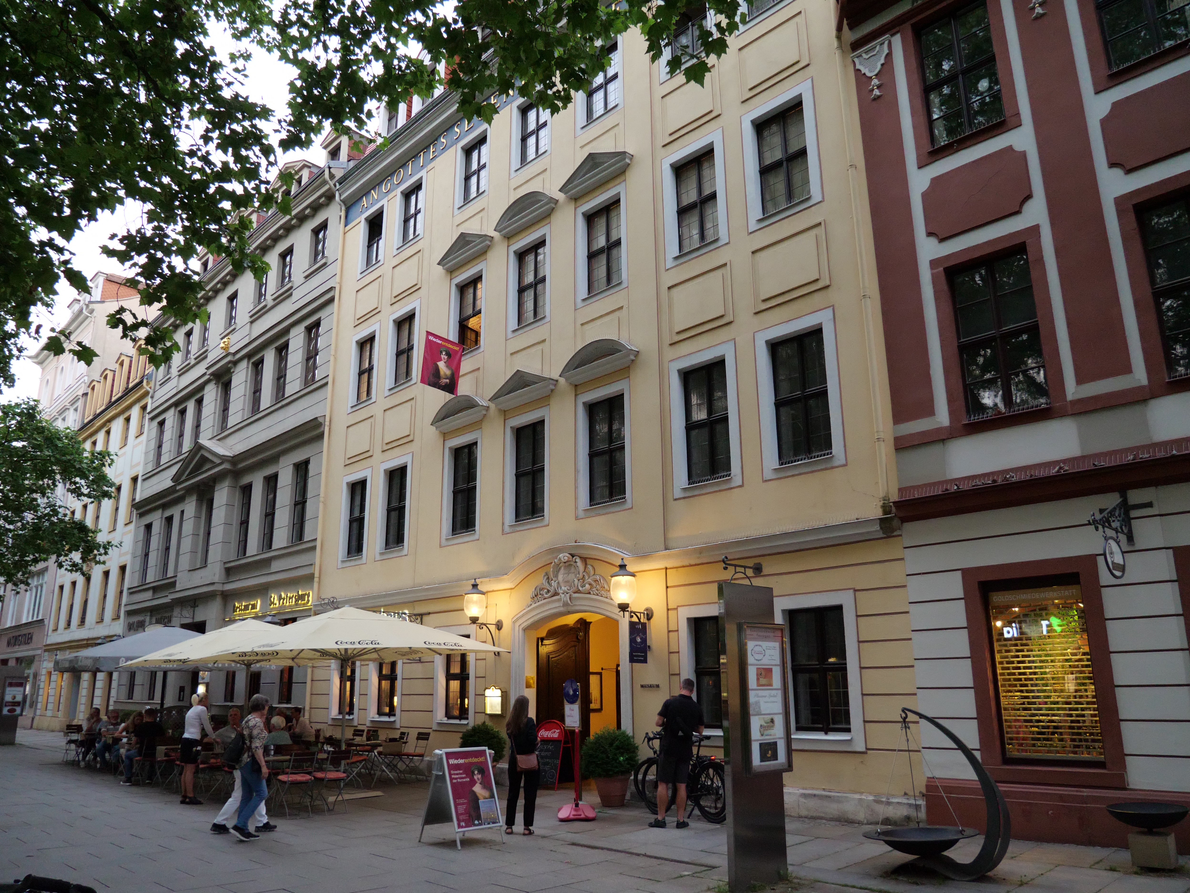 KI generiert: Das Bild zeigt eine Straße mit einem mehrstöckigen Gebäude im klassischen Stil, das ein Restaurant mit einem Außenbereich beherbergt. Menschen sitzen an Tischen unter Sonnenschirmen und gehen vorbei.