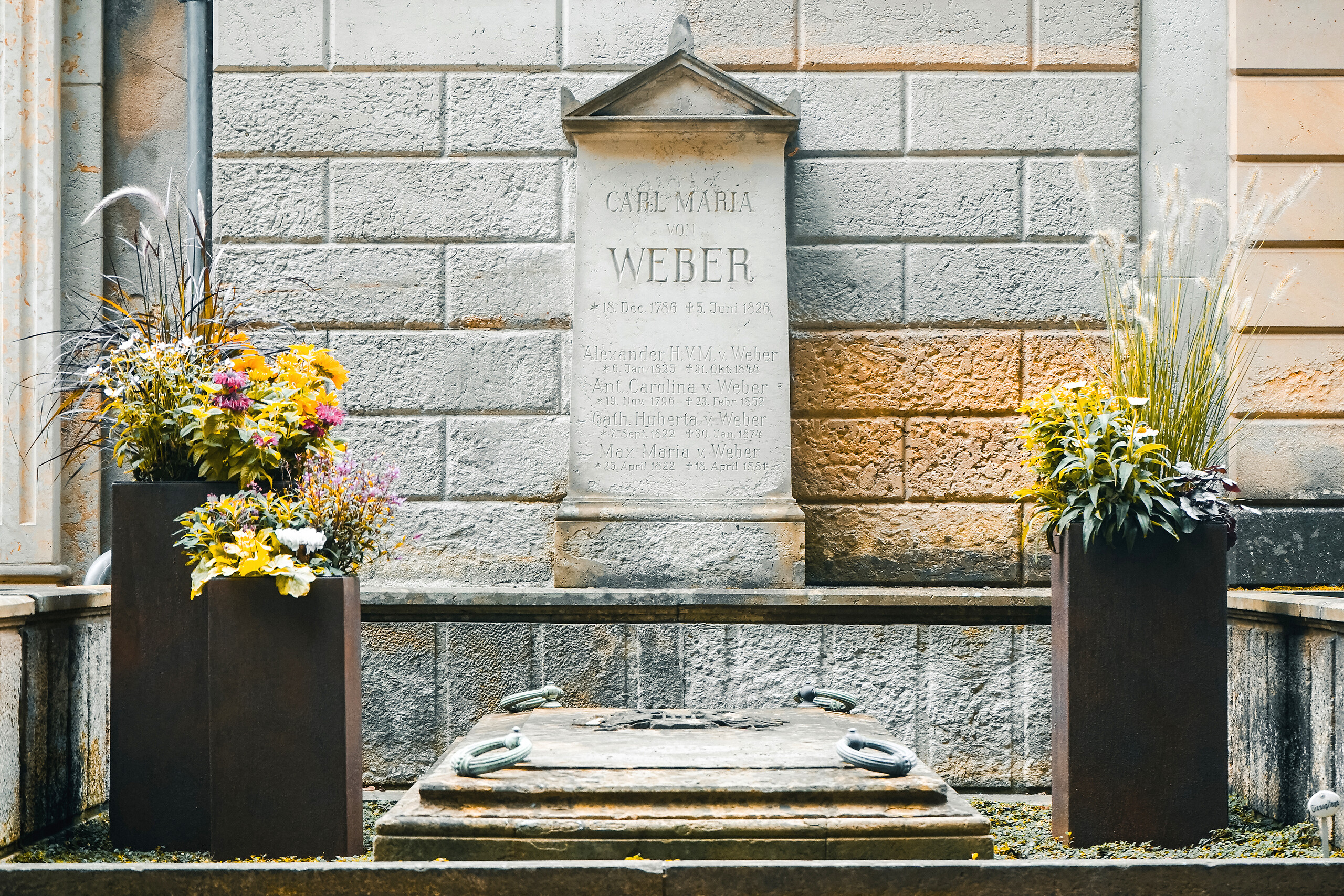 KI generiert: Das Bild zeigt ein Grabmal mit der Aufschrift "Carl Maria von Weber" und mehreren Blumengestecken. Es ist in eine Mauer integriert und wirkt gepflegt und respektvoll dekoriert.