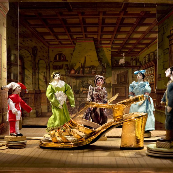 KI generiert: Das Bild zeigt eine inszenierte Szene mit kostümierten Puppen in einem historischen Raum, wobei ein großer goldener Schuh im Vordergrund steht. Die Figuren scheinen in einem Theaterstück oder einer künstlerischen Darstellung zu agieren.