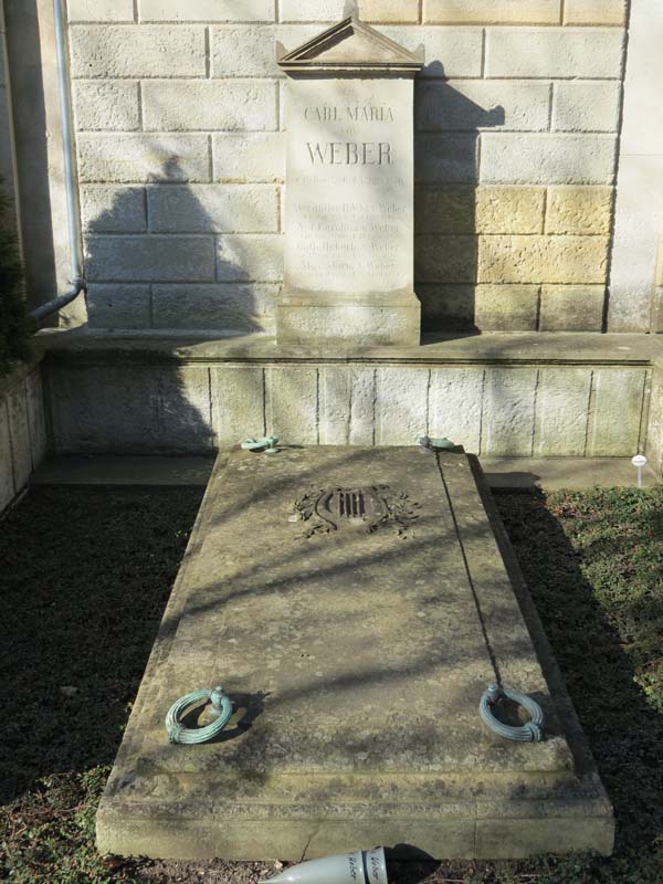 KI generiert: Das Bild zeigt einen Grabstein aus Stein, der an eine Mauer lehnt. Vor dem Stein liegt eine rechteckige Grabsplatte mit dekorativen Elementen.