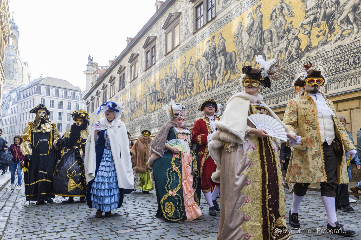 KI generiert: Das Bild zeigt eine Gruppe von Menschen in historischen Kostümen und Masken, die auf einer Kopfsteinpflasterstraße in einer Stadt spazieren gehen. Im Hintergrund ist ein großes Wandgemälde mit Reiterszenen zu sehen.